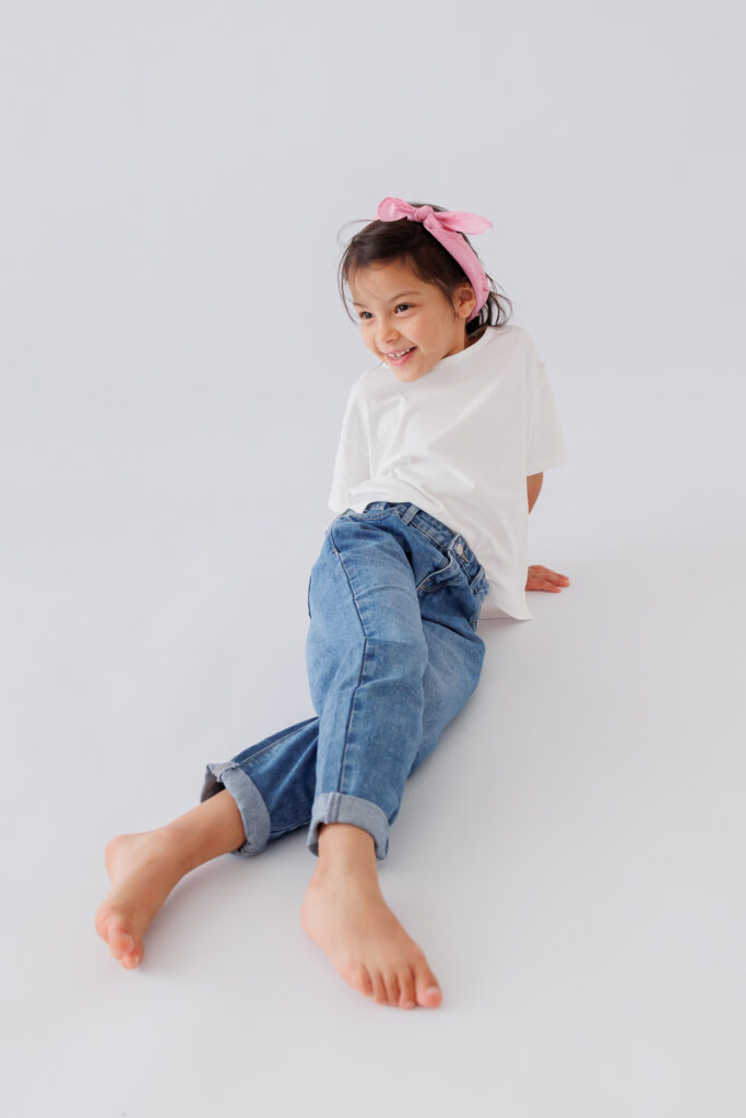 Mädchen sitzt lächelnd auf dem Boden in Jeans mit Haarband, Back-to-School-Mini-Session im Fotostudio Atelier 95 in Luxemburg