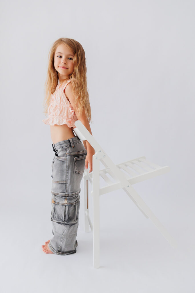 Mädchen lehnt sich seitlich an einen weißen Stuhl, rosa Top und graue Hose, Back-to-School-Mini-Session im Fotostudio Atelier 95 in Luxemburg